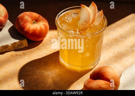 Ein erfrischendes Glas Pfirsicheistee mit Eiswürfeln und frischen Pfirsichscheiben, die das Getränk garnieren, vor einer strukturierten Steinoberfläche und umgeben von B Stockfoto