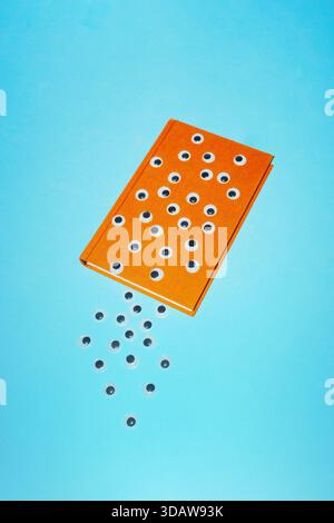 Ein oranges Buch, das kreativ mit zahlreichen Googly-Augen verziert ist, liegt auf einer hellblauen Oberfläche, mit einigen Augen, die eine skurrile Darstellung erzeugen Stockfoto