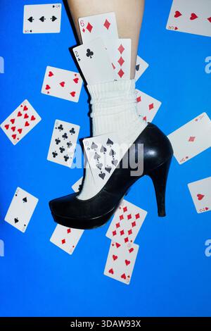 Konzeptionelles Modebild, das das Bein einer Frau mit schwarzem High Heel und weißer Socke zeigt, mit Spielkarten, die auf einem bo verstreut sind Stockfoto