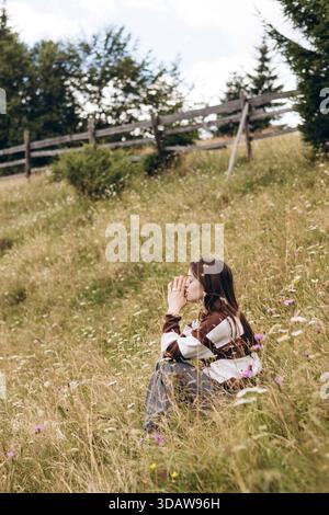 Eine junge Frau sitzt friedlich inmitten von Wildblumen auf einem grasbewachsenen Hügel, umgeben von malerischen Bergen und Natur, und genießt die Ruhe und frische A Stockfoto
