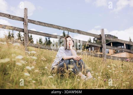 Eine junge Frau sitzt friedlich auf einer grasbewachsenen Wiese, umgeben von Bergen und einem rustikalen Holzzaun und genießt an klaren Tagen die Ruhe der Natur Stockfoto