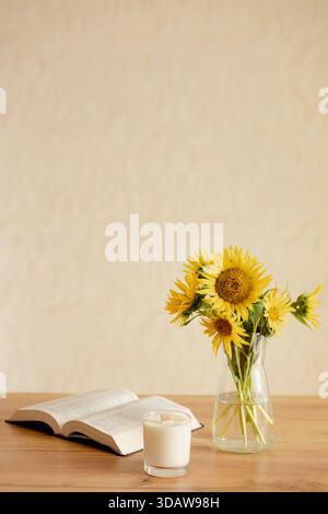 Ein ruhiges Stillleben mit leuchtenden Sonnenblumen in einer klaren Vase, begleitet von einem offenen Buch und einer beleuchteten Kerze auf einem Holztisch in einer gemütlichen Ho Stockfoto