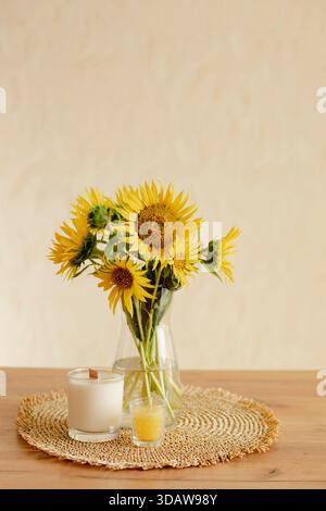 Ein bezauberndes Stillleben mit einem leuchtenden Sonnenblumenstrauß in einer klaren Vase mit zwei Kerzen, die in der Nähe auf einer gewebten Matte platziert werden. Es bringt Wärme in ein gemütliches Stockfoto