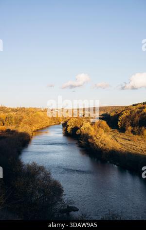 Ein ruhiger Fluss fließt durch eine malerische Herbstlandschaft. Goldenes Laub säumt die Flussufer und reflektiert die warmen Farben des Herbstes in klarem Blau Stockfoto