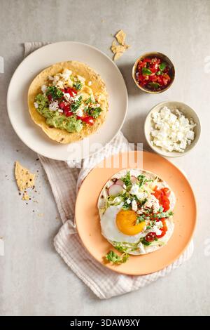 Blick von oben auf zwei Teller mit lebhaften Huevos Rancheros auf Tortillas. Eine mit Spiegelei, Tomate, Avocado und Koriander auf einem Orangenteller und eine andere Stockfoto