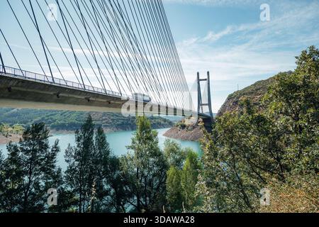 Eine moderne, mit Kabel gehaltene Brücke überspannt einen ruhigen Wasserweg, umgeben von üppigen grünen Hügeln und Bäumen. Der klare blaue Himmel trägt zur Ruhe und Landschaft bei Stockfoto