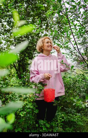 Eine Frau in einem rosafarbenen Hoodie erntet reife Früchte in einem grünen Garten. Sie steht inmitten von üppigem Grün, hält einen roten Korb, umhüllt die Ruhe Stockfoto