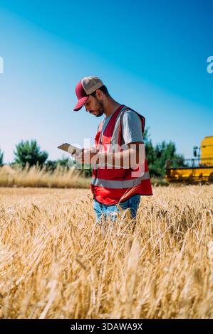 Ein Landwirt in Guadalajara, Spanien, verwendet ein digitales Tablet, um Erntedaten auf einem Gerstenfeld zu überwachen. Der blaue Himmel und Mähdrescher im Hintergrund hig Stockfoto