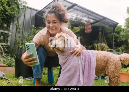 Eine fröhliche Frau nimmt mit ihrem Smartphone einen Moment mit ihrem Hund in ein Handtuch gewickelt ein und genießt einen sonnigen Tag in ihrem üppigen Garten in Australien. Stockfoto
