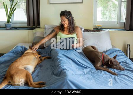 Eine Frau genießt einen ruhigen Morgen im Bett, umgeben von zwei schlafenden Hunden. Sie lächelt warm, während sie eine Tafel hält und die Ruhe und Begleiter umschließt Stockfoto