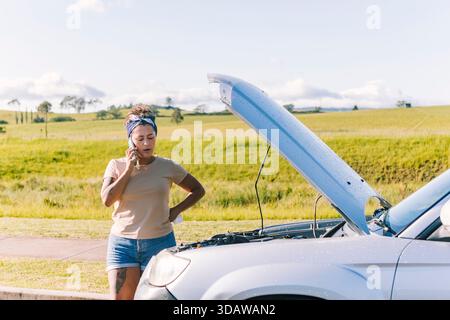 Eine Frau, die neben ihrem kaputten Auto auf einer Landstraße in Australien steht und ein Telefon hält. Die offene Motorhaube und die malerische Landschaft betonen die Isolat Stockfoto