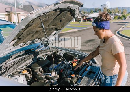 Eine Frau steht an einem sonnigen Tag in einem australischen Vorort neben ihrem kaputten Auto, inspiziert den Motor unter der geöffneten Motorhaube und denkt über die nächsten Schritte nach Stockfoto