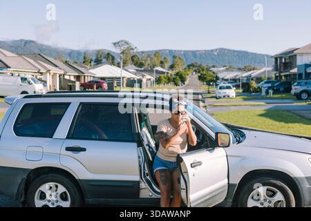 Eine Frau steht neben ihrem kaputten Auto auf einer australischen Vorstadtstraße und sucht Hilfe. Der klare Himmel und die Wohngegend machen das deutlich Stockfoto