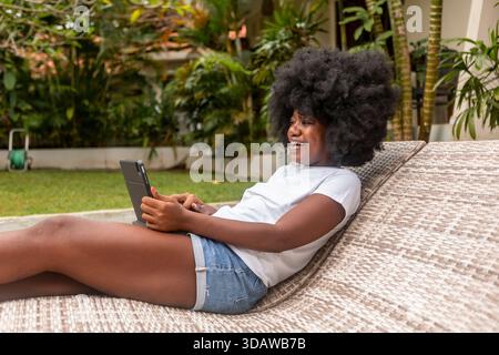 Afroamerikanerin mit Afro-Frisur, die als digitaler Nomade auf Bali fernarbeitet. Entspannt und lächelnd, während Sie ein Tablet verwenden, umgeben von lu Stockfoto