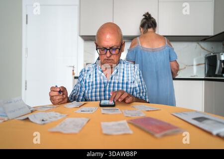 Ein älterer Mann, der die Haushaltsfinanzen verwaltet, mit Quittungen auf dem Tisch. Er benutzt einen Taschenrechner auf einem Smartphone, während eine nicht erkennbare Frau in der Bank ist Stockfoto
