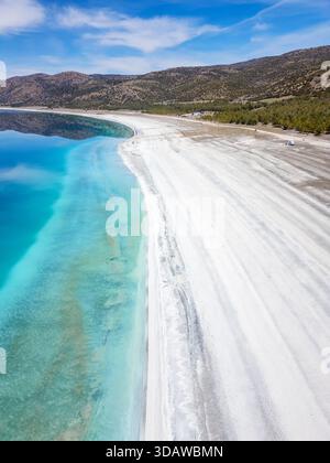 Aus der Vogelperspektive auf das unberührte türkisfarbene Wasser des Salda Crater Lake trifft auf die hellen weißen Sandstrände, umgeben von üppigem Grün und fernen Hügeln unter Stockfoto
