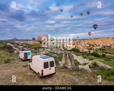 Aus der Vogelperspektive auf die im Love Valley in Kappadokien geparkten Wohnmobile, umgeben von farbenfrohen Heißluftballons, die am ruhigen Morgen schwimmen Stockfoto