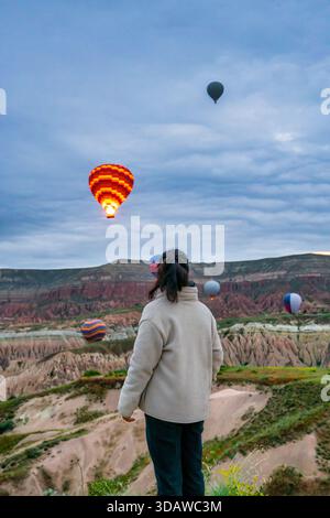 Eine Frau blickt auf lebendige Heißluftballons, die über Kappadokiens atemberaubende Täler schweben und ein atemberaubendes Panorama aus bunten Ballons gegen dr. Stockfoto