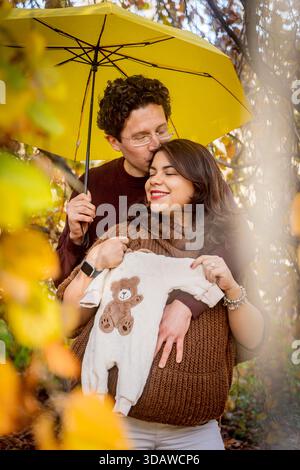 Ein liebevolles Paar feiert die Schwangerschaft in einer wunderschönen Herbstkulisse. Der Mann küsst die Frau liebevoll unter einem gelben Regenschirm, während sie ein Baby hält Stockfoto
