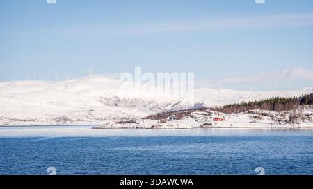 Schneebedeckte Landschaft in Kvaloysletta in Norwegen mit einer Sammlung moderner Windturbinen auf den Hügeln, mit kristallklarem blauem Wasser im Vordergrund Stockfoto