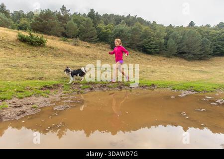 Eine Frau in lebendiger sportlicher Ausrüstung macht einen Trail-Lauf neben ihrem Hund. Sie bewegen sich auf einem landschaftlich reizvollen Outdoor-Pfad und spiegeln aktive Lebensstile und Begleitpersonen wider Stockfoto