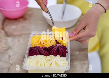 Eine Nahaufnahme einer Hand, die leuchtendes gelbes, lila und cremefarbenes Eis aus einem Behälter schöpft. Die Texturen des Eiss werden in t hervorgehoben Stockfoto