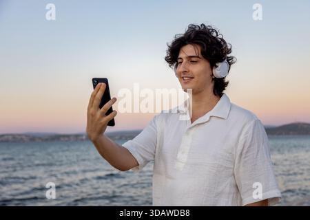 Ein junger Mann mit Kopfhörern lächelt, während er ein Handy hält, am Meer steht. Der Sonnenuntergang färbt den Himmel mit sanften Farben und schafft eine ruhige Atmosphäre Stockfoto