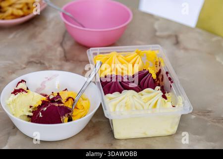 Eine Nahaufnahme von bunten Eiskugeln in einer weißen Schüssel und Tablett auf einem Marmortisch. Löffel werden in beide Schalen gelegt, um die Textur und Crea zu betonen Stockfoto