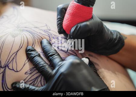 Eine Tätowierungskünstlerin färbt geschickt ein detailliertes Blumendesign auf die Haut eines Kunden. Das Bild fängt Präzision und Kunstfertigkeit ein und hebt das kreative hervor Stockfoto
