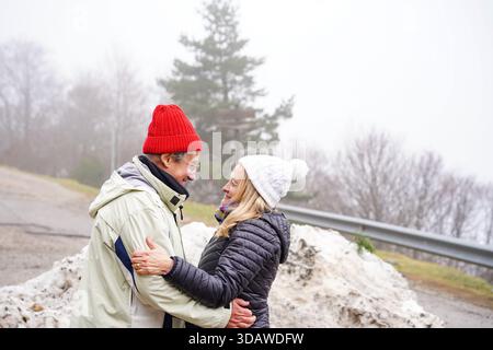 Ein älteres Paar wird bei einem Winterausflug fröhlich gefangen gehalten, warm gekleidet mit Hüten und Jacken, umgeben von einer malerischen Schneelandschaft Stockfoto