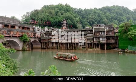 Ein traditionelles Holzboot gleitet entlang des Flusses in Richtung Phoenix Hong Bridge, wo die malerische Stelzen Building Group wunderschön eingerahmt ist Stockfoto