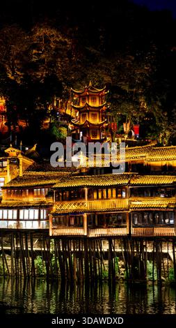 Ein atemberaubender Blick bei Nacht auf die Altstadt von Fenghuang, China, mit traditionellen Stelzengebäuden und einer wunderschön beleuchteten Pagode. Die Reflexion leuchtet auf Stockfoto