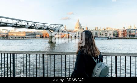Rückansicht einer unerkennbaren Frau, die an der Themse steht und auf die Millennium Bridge und St Paul's Cathedral blickt. Ein ruhiger Moment, der Londons Wahrzeichen festhält Stockfoto