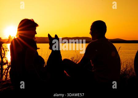 Eine Familie sitzt am ruhigen See mit ihrem Hund und genießt einen faszinierenden Sommersonnenuntergang. Die warmen Töne des Himmels spiegeln sich auf dem Wasser und bilden eine Pässe Stockfoto