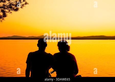 Ein Paar hat eine Silhouette vor den lebhaften Farben eines Sonnenuntergangs an einem ruhigen See, was eine ruhige und romantische Atmosphäre schafft, während der Tag in den Tag übergeht Stockfoto
