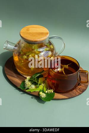 Von oben werden eine Glas-Teekanne und eine Tasse gefüllt mit beruhigendem Kräuterlindentee auf ein rundes Holztablett gestellt. Frische Lindenblüten umgeben die Einrichtung o Stockfoto