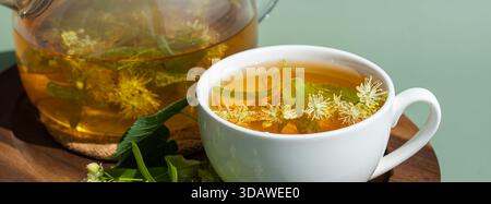 Eine wohltuende Tasse Kräuterlindentee mit frischen Lindenblüten in einer weißen Tasse. Eine transparente Teekanne mit mehr Lindenaufguss befindet sich auf einem Holz Stockfoto