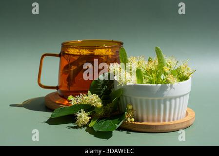 Eine beruhigende Szene mit Kräuterlindentee in einem bernsteinfarbenen Glasbecher neben frischen Lindenblüten in einer weißen Keramikschale auf einem hölzernen Untersetzer Stockfoto