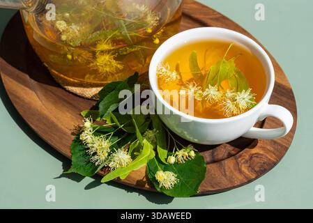 Von oben liegt eine weiße Tasse Kräuterlindentee auf einem Holztablett, umgeben von frischen Lindenblüten. Der sanfte grüne Hintergrund verstärkt den Tee Stockfoto