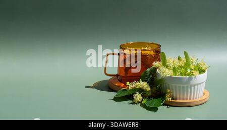 Eine warme Bernsteinglasschale mit Kräuterlindentee steht auf einem hölzernen Untersetzer, umgeben von frischen Lindenblüten in einer weißen Schüssel auf einem sanften grünen Rücken Stockfoto
