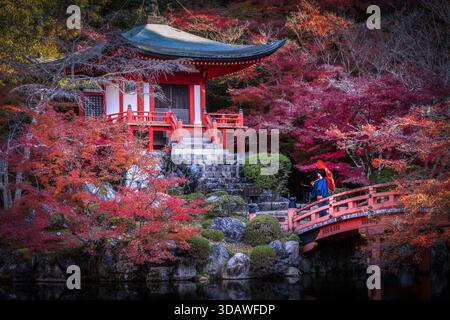 Der Blick auf die lebendigen Ahornbäume umrahmt eine traditionelle rot-weiße Pagode, während eine Frau mit einem roten Regenschirm über eine Brücke in Kyoto, Kyoto, Japan überquert. Stockfoto