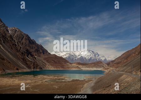 Der Blick auf das türkisfarbene Wasser trifft auf zerklüftete, von Schnee gekrönte Berge unter hellem Himmel, gefangen in Sadpara, Skardu, Pakistan. Stockfoto