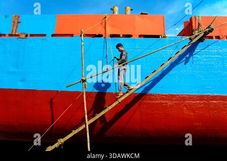 Dhaka, Bangladesch - 31. August 2024: Blick auf einen Arbeiter, der auf einer Bambusleiter unsicher balanciert ist und den leuchtenden blauen und roten Rumpf eines großen Schiffes malte Stockfoto