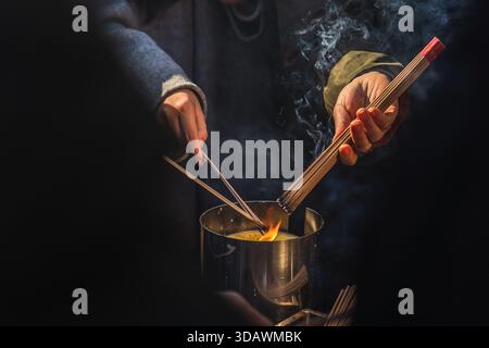 Peking, China - 26. November 2025: Blick auf die Hände, die vorsichtig Weihrauchstäbchen anzünden, Rauch kräuselt sich in einem rituellen Tanz gegen einen harten, schattigen Schattenwurf nach oben Stockfoto