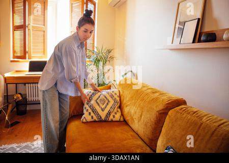 Das weibliche Individuum passt ein gemustertes Kissen auf einem stilvollen Sofa in einem gut beleuchteten Wohnzimmer an, umgeben von Grün und einer warmen Atmosphäre Stockfoto