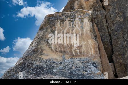 Antike gobustanische Petroglyphen tanzender menschlicher Figuren, die auf einer zerklüfteten Felsoberfläche unter blauem Himmel mit Wolken in Aserbaidschan geätzt sind. Stockfoto