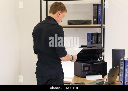 Der Mann hält Dokumente vom Drucker aus in einem Arbeitsbereich mit Regalen und Ordnern Stockfoto