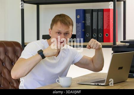 Ein aufgeregter junger Büroangestellter lächelt und beugt beide Arme in Siegerposition am Schreibtisch mit Laptop, Kaffeetasse und Aktenmappen in der Nähe. Stockfoto
