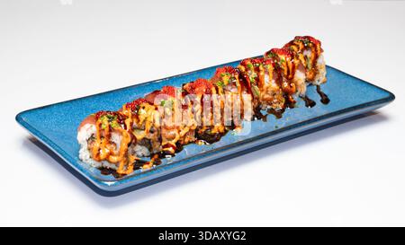 Scharfe Sushi-Brötchen mit Lachs, Reis und Gemüse, garniert mit cremigen und dunklen Saucen und Fischrogen, serviert auf einer blauen Keramikplatte isoliert auf weiß Stockfoto