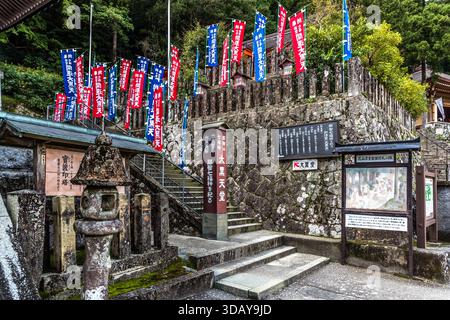 Farbenfrohe Flaggen wehen am Eingang zur Kumano Kodo-Pilgerroute vor einer Steinmauer in Nachikatsuura, Präfektur Wakayama, Japan. Stockfoto